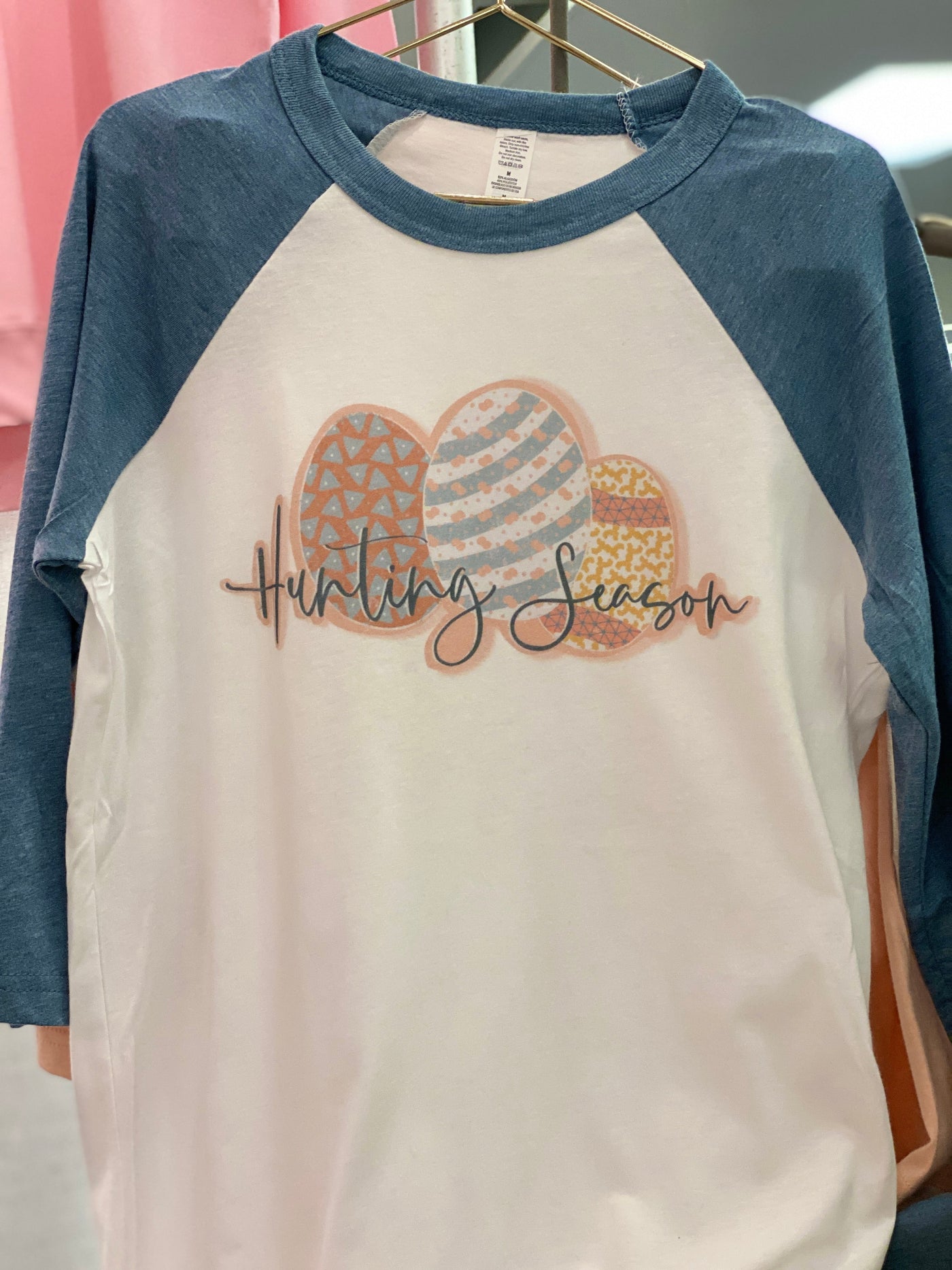 A white raglan with blue sleeves and a graphic of three differently decorated easter eggs and the words "Hunting Season" In a blue cursive font