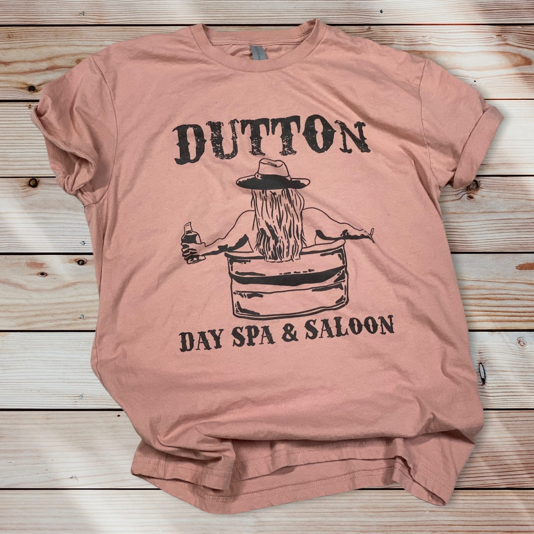 Pink shirt with a black graphic on it. The graphic Has the word Dutton in old western letters at the top and Day Spa & Saloon at the bottom, in between the words you can see a womans back with long hair under her cowboy hat sitting in a water trough holding a bottle of whiskey.
