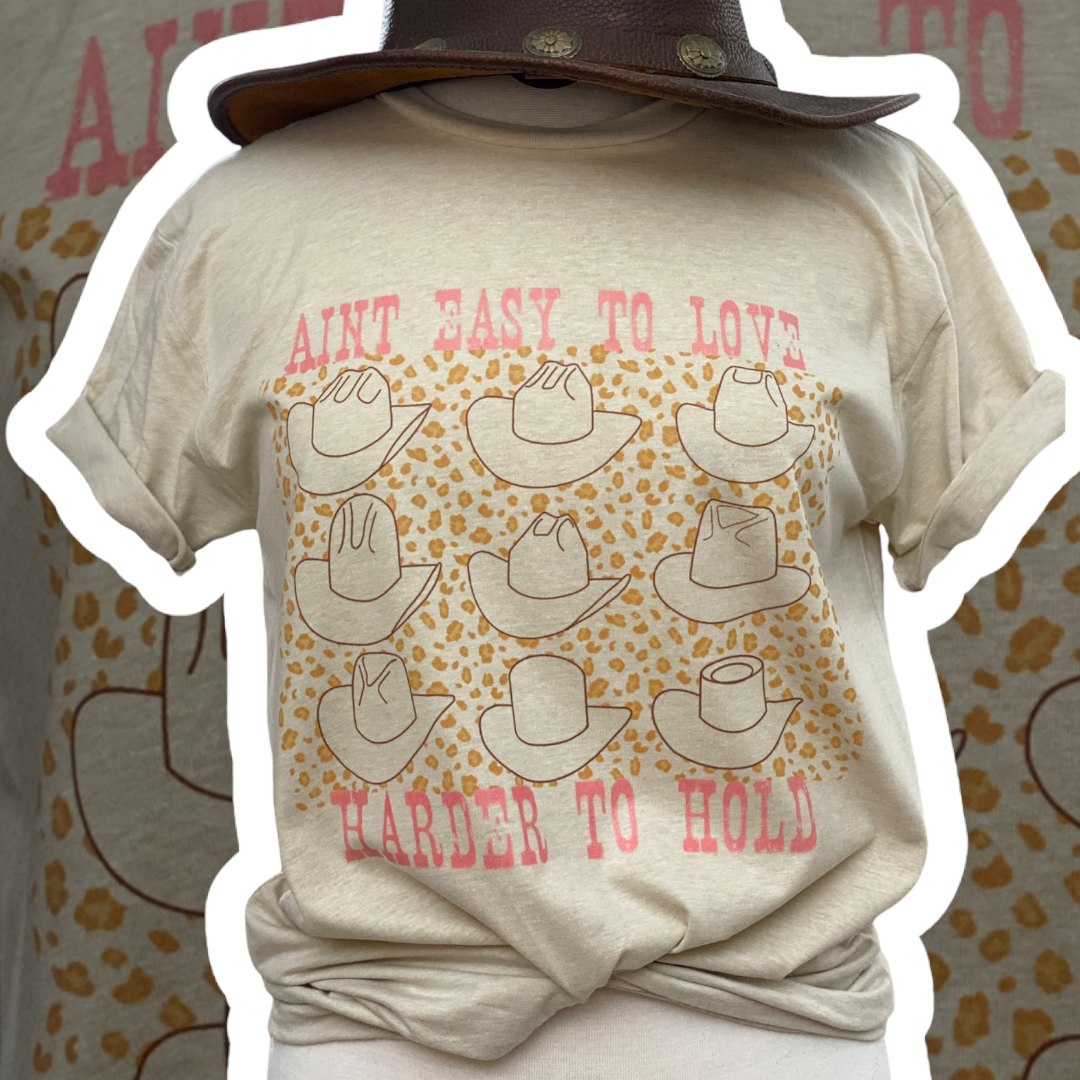 Tan tee with graphic. Graphic is pink weathered letters at the top that read " Aint Easy To Love" underneath the text there are 9 different styles of cowboy hats outlined in brown with a light brown leopard print in between the cowboy hats, text below the hats reads " Harder To Hold"