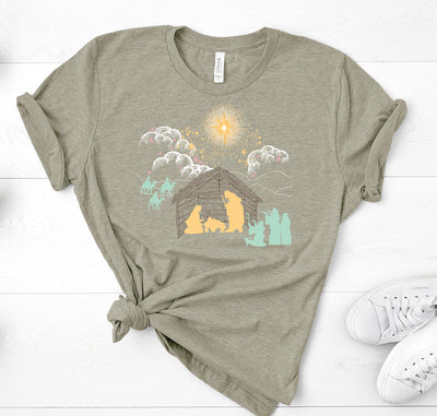 Heather Olive Green T Shirt depicting a nativity scene. White clouds Gold Stars. Yellow Mary Joseph and baby Jesus silhouette, all other silhouettes are light teal  