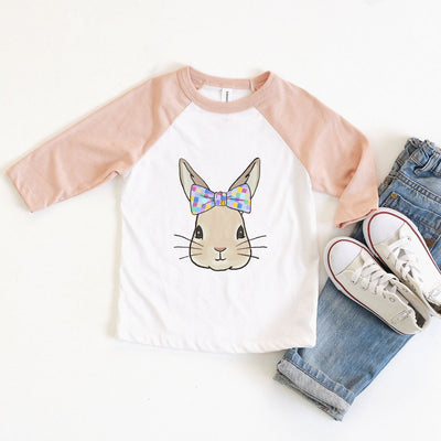 a white raglan with peach sleeves and the graphic of a bunny wearing a bow with multicolored squares on it.