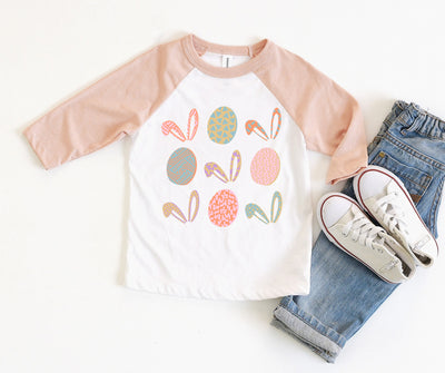 a white raglan tee with peach sleeves and a graphic of tic tac toe of different colored and patterned easter eggs and bunny ears