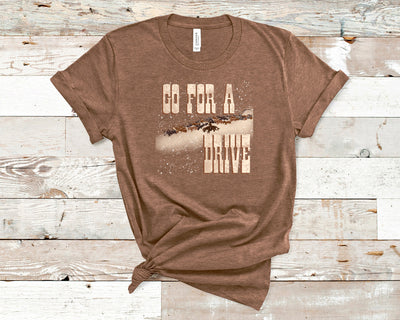 A heather brown graphic tee. The graphic is of cowboys on a cattle drive with the words "Go for a drive" in a western font. there are white specks over the words and art to emulate snow. 