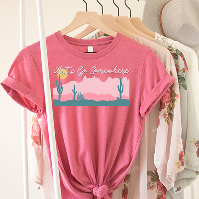 Heather Pink Shirt with graphic. Graphic is of a desert scene. Foreground is turquoise withe a dark pink as the mid ground and light pink on the horizon and a yellow sun. On top of the graphic are the words Let's Go Somewhere in a white cursive font with a  turquoise shadow. 