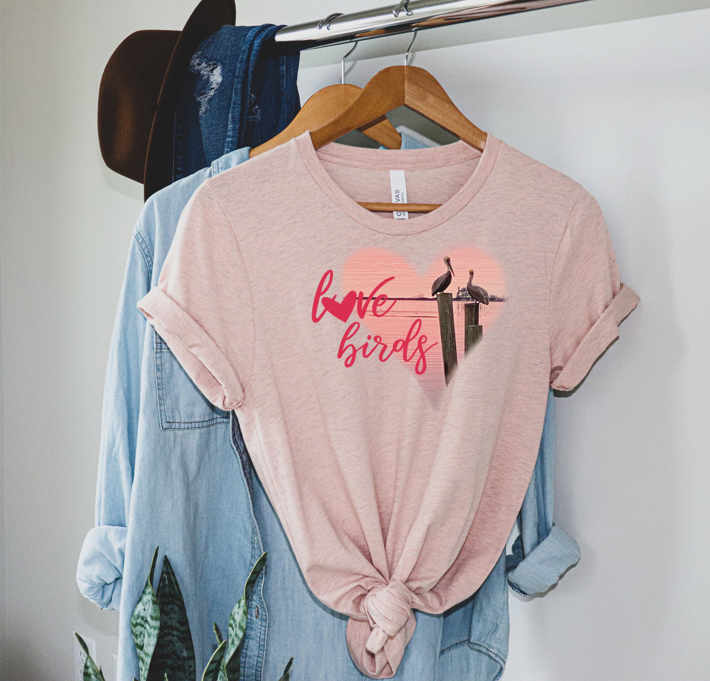 Heather peach tee with graphic. Graphic is a pink heart with a photo of some pelicans standing on post in the water. To the left of the heart are the words love birds in a pink cursive font and the O in love has been replaced with a heart.