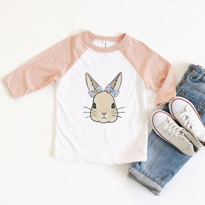 A white raglan tee with peach sleeves and a graphic of a bunny wearing a bow with multicolored squares on each ear 