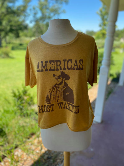 Mustard yellow shirt. Graphic says Americas most wanted with a photo of Rip from Yellowstone in brown.
